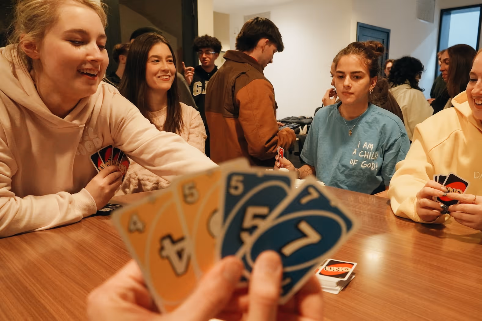 Group of young adults playing UNO card game around a wooden table indoors.
