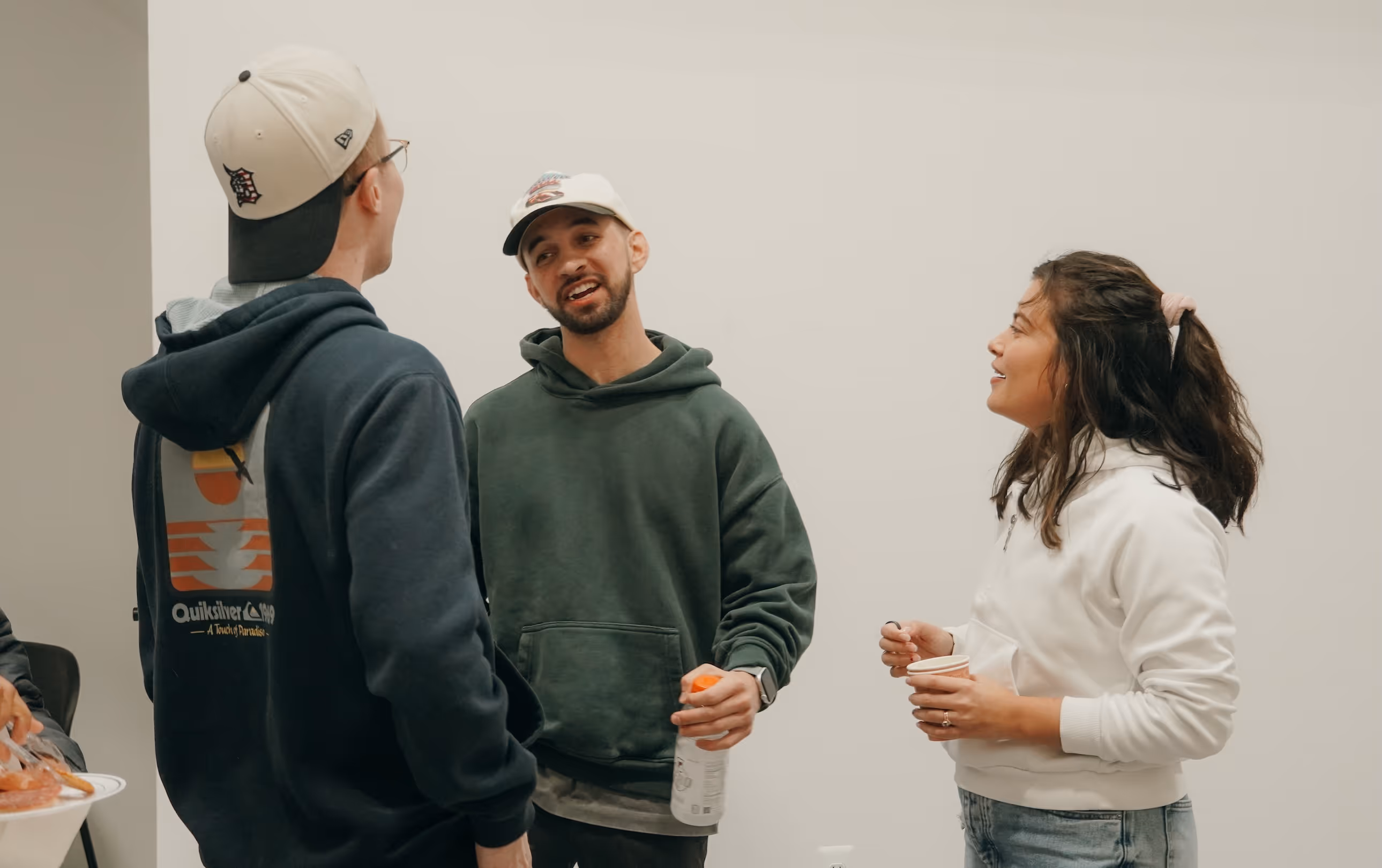 Three young adults standing and talking indoors, two men wearing caps and hoodies, one woman in a white hoodie holding a cup.