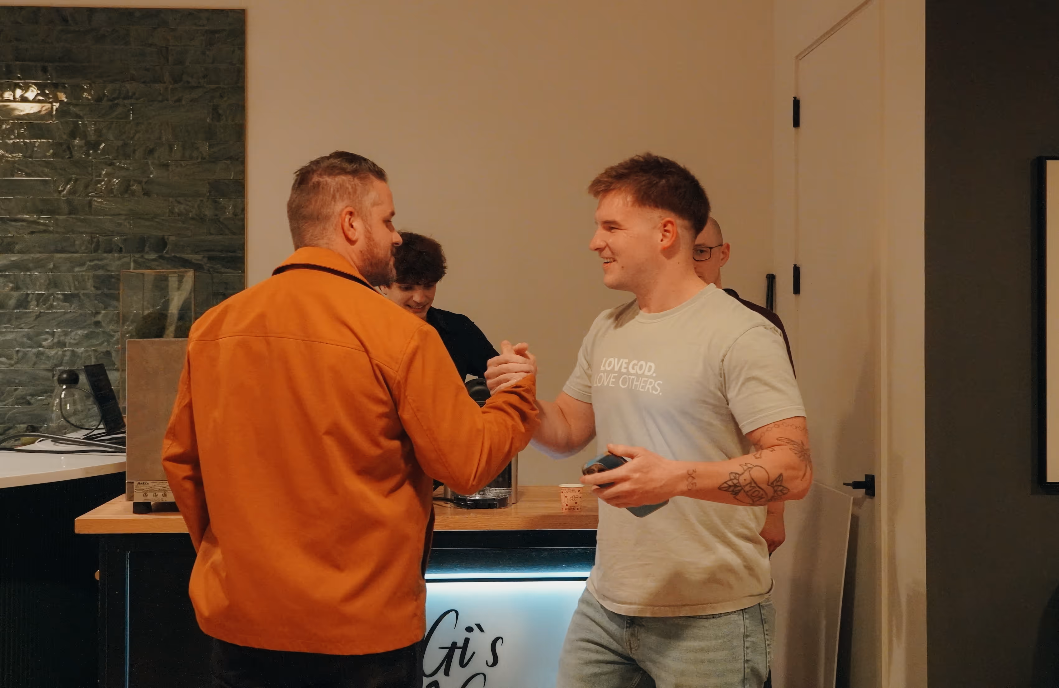 Two young men smiling and shaking hands in a casual indoor setting with others in the background.