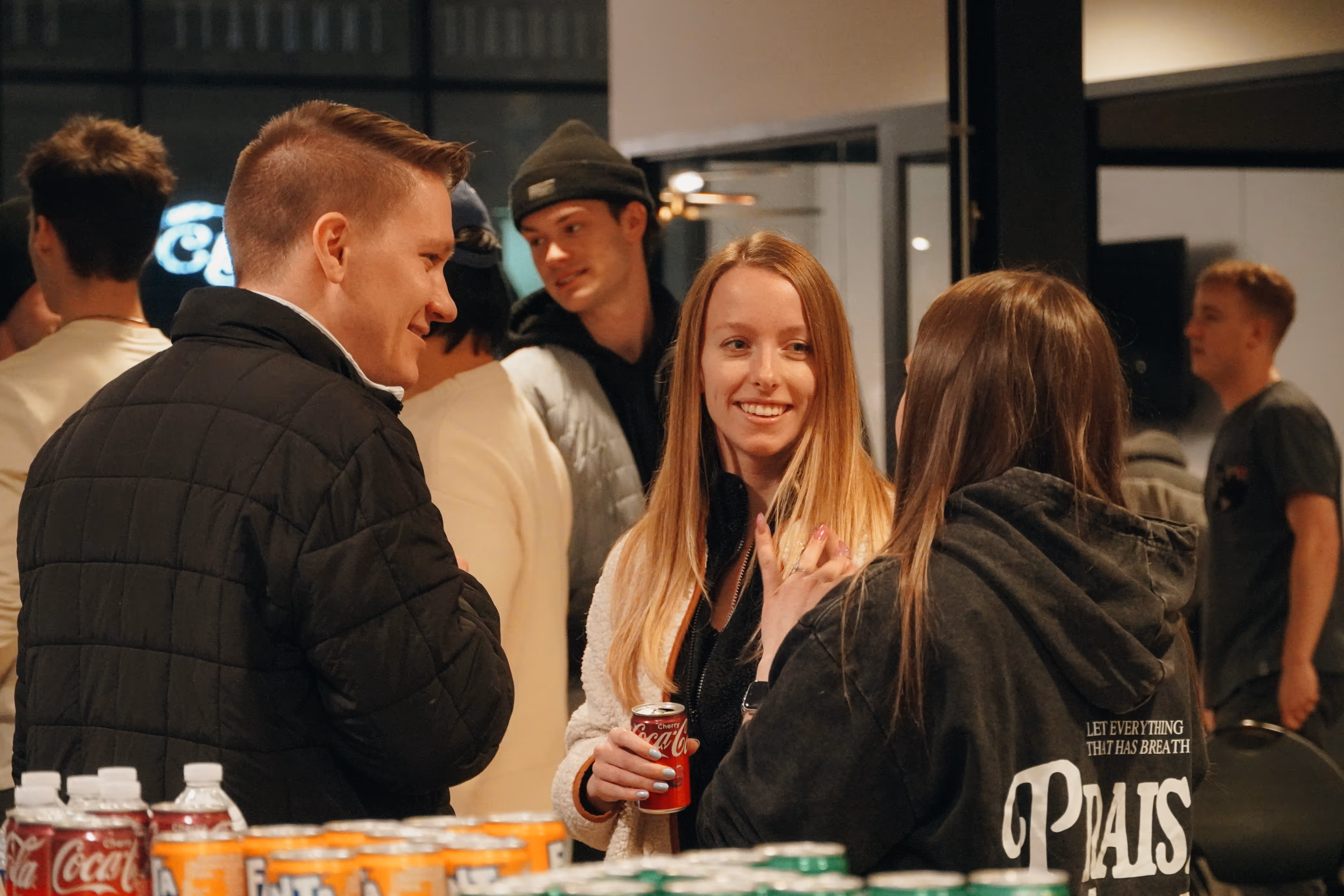 Three young adults casually chatting indoors, one holding a can of cherry Coca-Cola, with soda cans visible in the foreground.