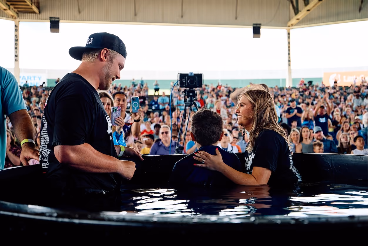 Man and woman baptize a child in a water tank with a crowd watching in the background.
