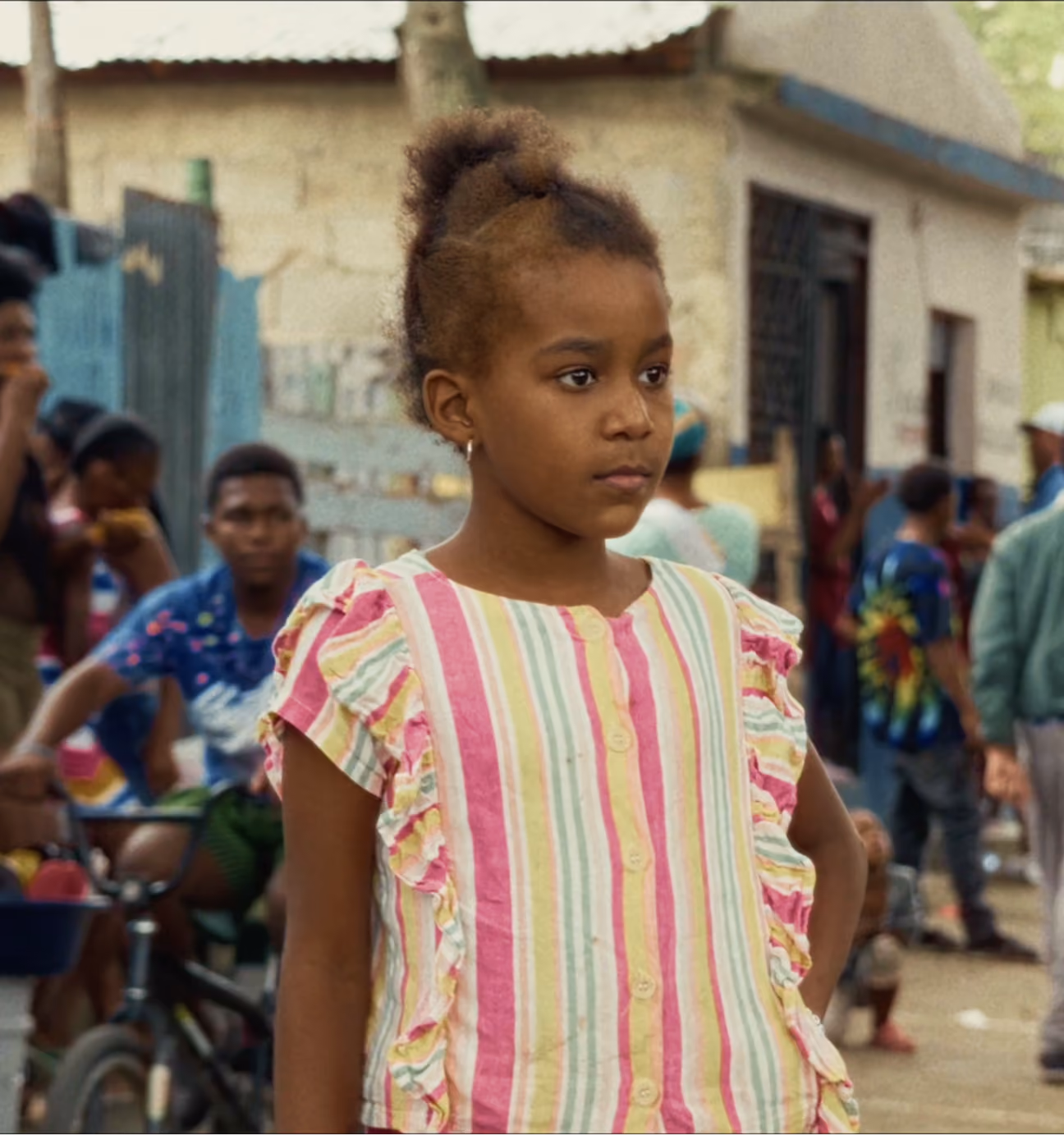 Young girl in a pink and yellow striped dress standing with a serious expression outdoors.