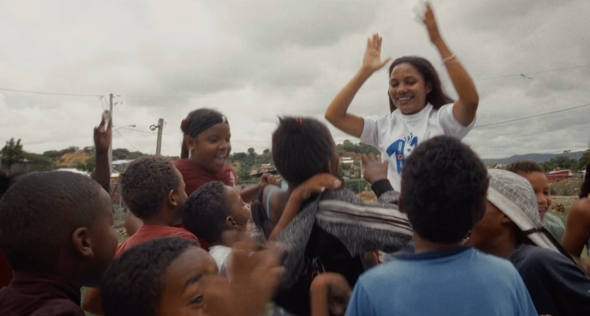 Group of children joyfully playing outside with a woman lifting her arms and smiling.