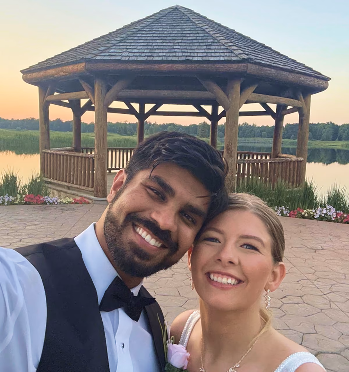 Smiling couple in formal wear taking a selfie outdoors near a wooden gazebo by a lake at sunset.
