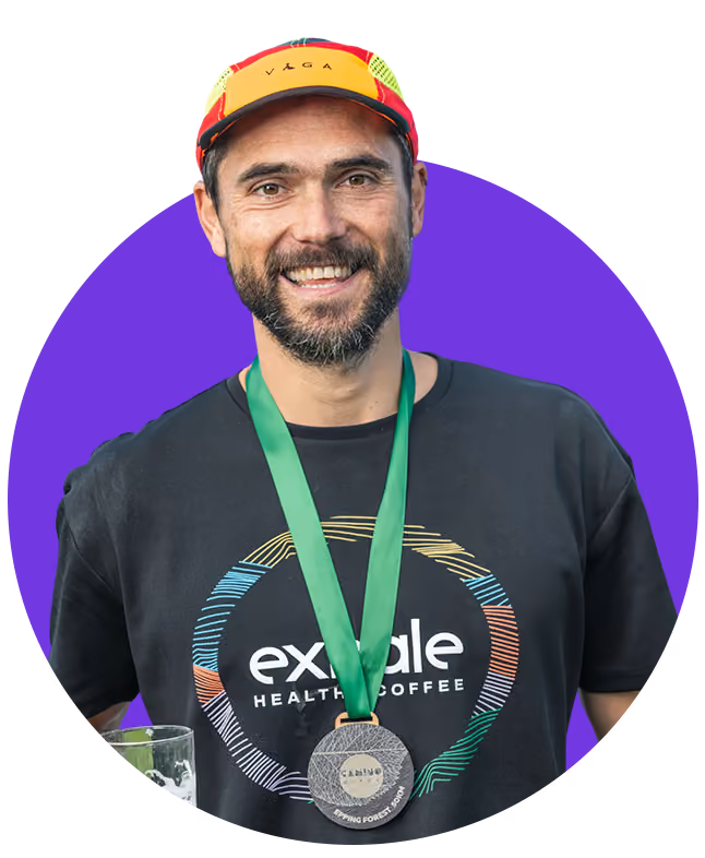 Smiling man wearing a colorful cap, a black 'exmale healthy coffee' t-shirt, and a medal around his neck holding a glass of beer.
