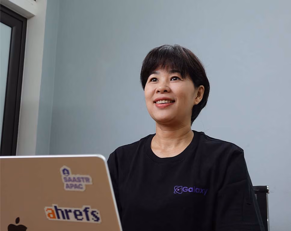 Smiling woman with short dark hair wearing a black Galaxy t-shirt sitting with a laptop stickers 'SAASTR APAC' and 'ahrefs'.