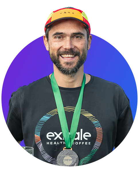 Smiling man wearing a red and yellow cap, black 'Exhale Healthy Coffee' t-shirt, and a green medal ribbon around his neck, holding a glass.