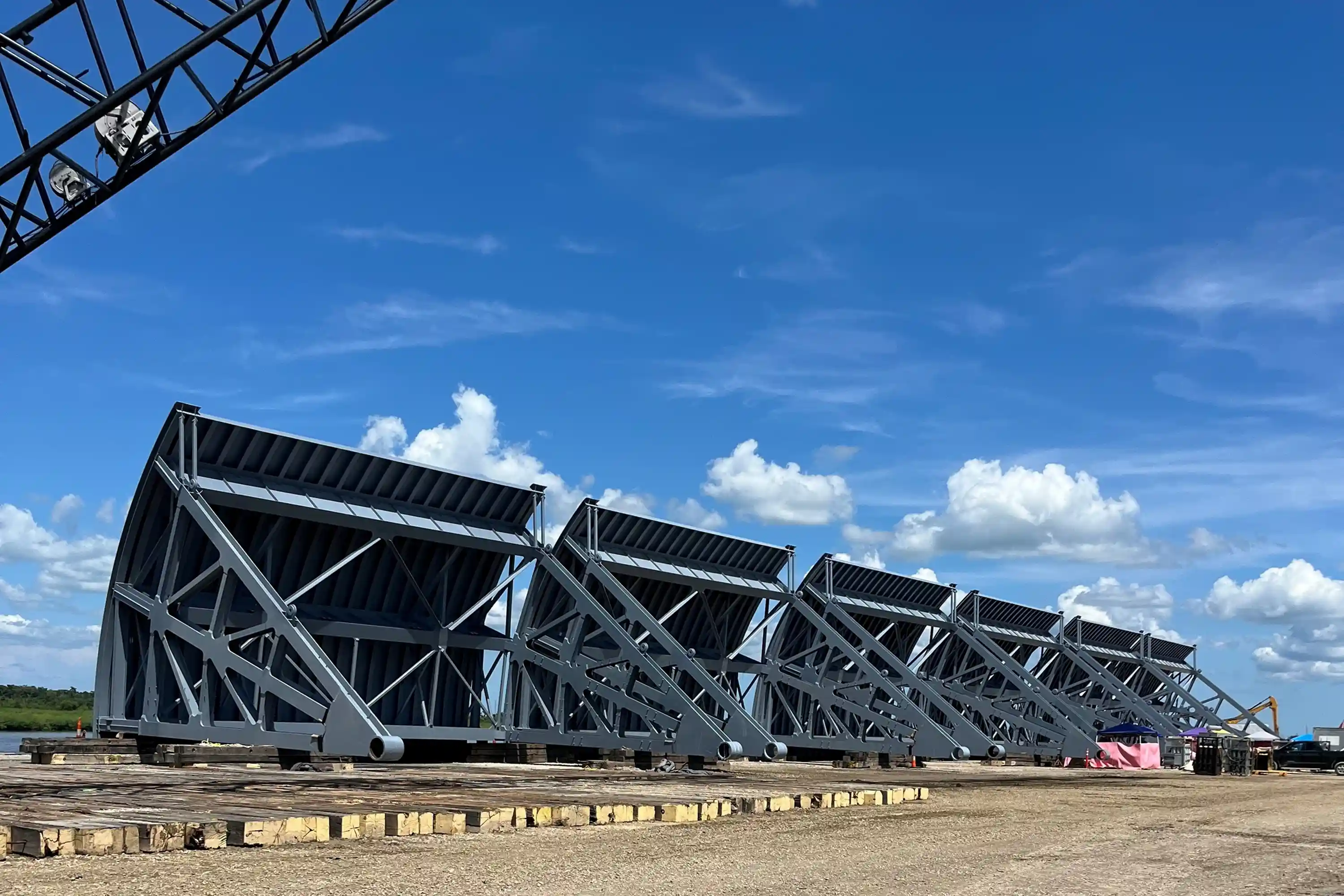 Large gray steel truss structures lined up on a gravel ground under a blue sky with scattered clouds.