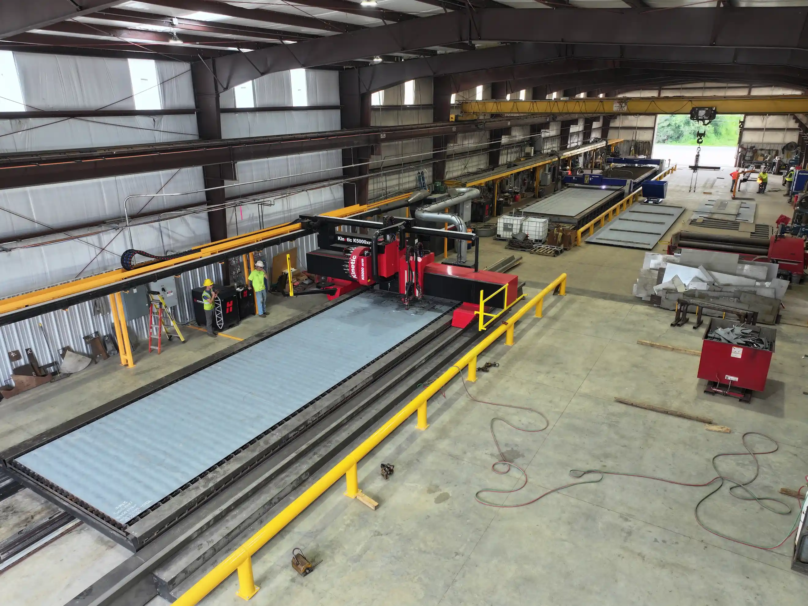 Large industrial warehouse with machinery and workers operating a red kinetic K5000XT cutting machine on a metal sheet.