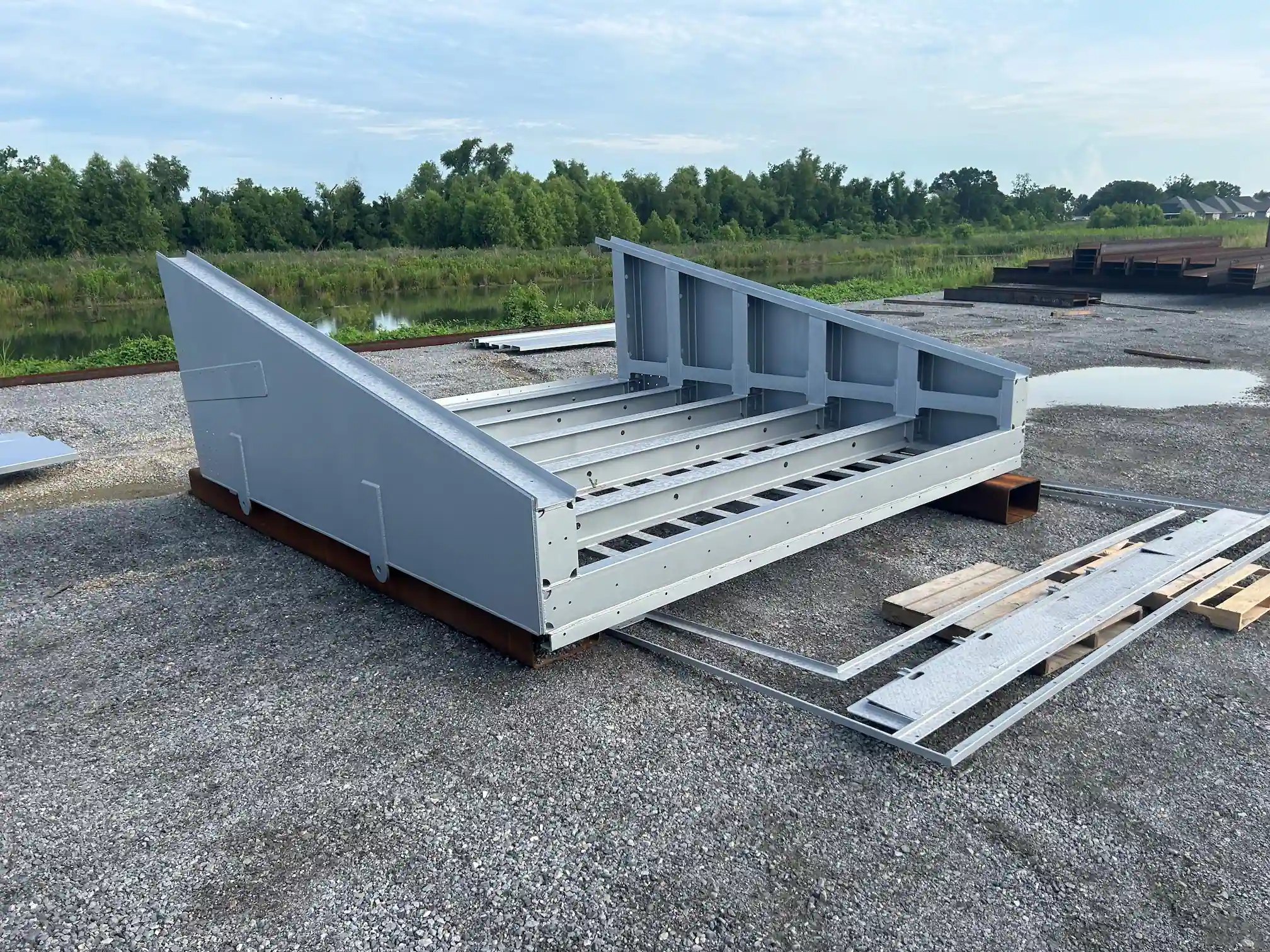 Large metal industrial frame or structure resting on gravel with green trees and a pond in the background.