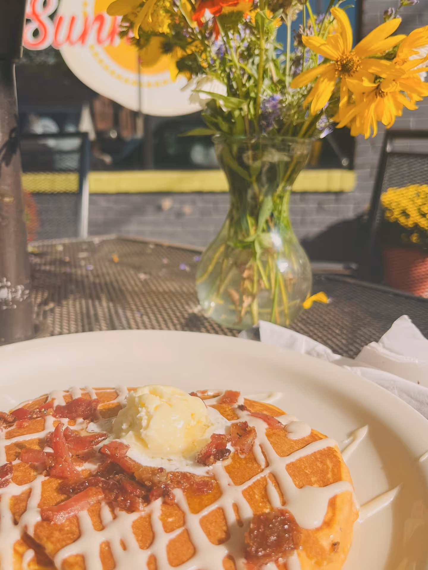 A waffle topped with bacon, butter, and cream drizzle sits on a white plate in sunlight. A vase of vibrant yellow flowers adds a cheerful touch.