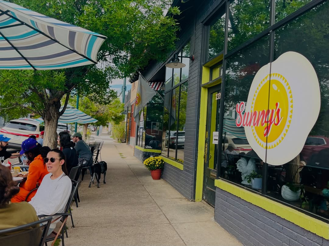 Outdoor cafe scene with people dining under striped umbrellas on a sidewalk. A large window displays “Sunny's” logo.