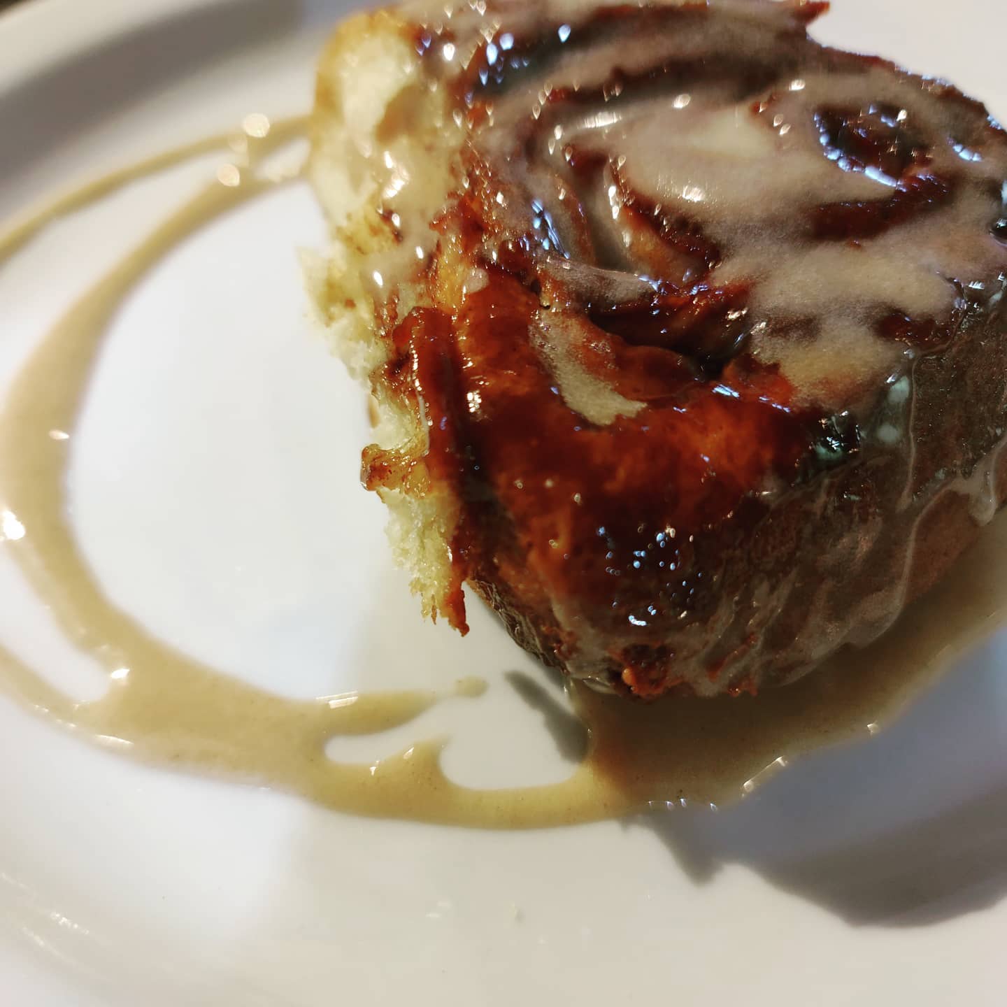 Close-up of a cinnamon roll on a white plate, drizzled with glossy icing. The roll has a golden-brown crust, evoking warmth and sweetness.