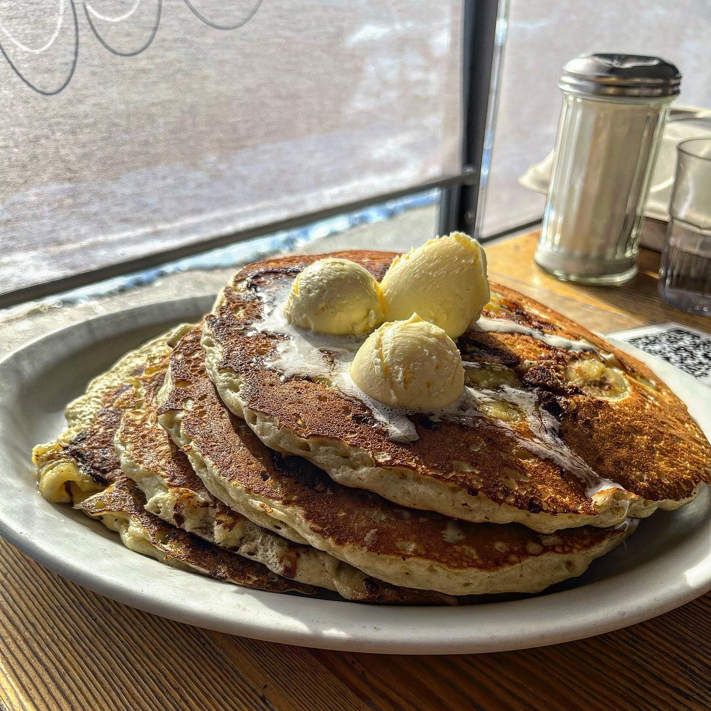 A stack of four golden-brown pancakes topped with melting butter sits on a white plate at a wooden table.