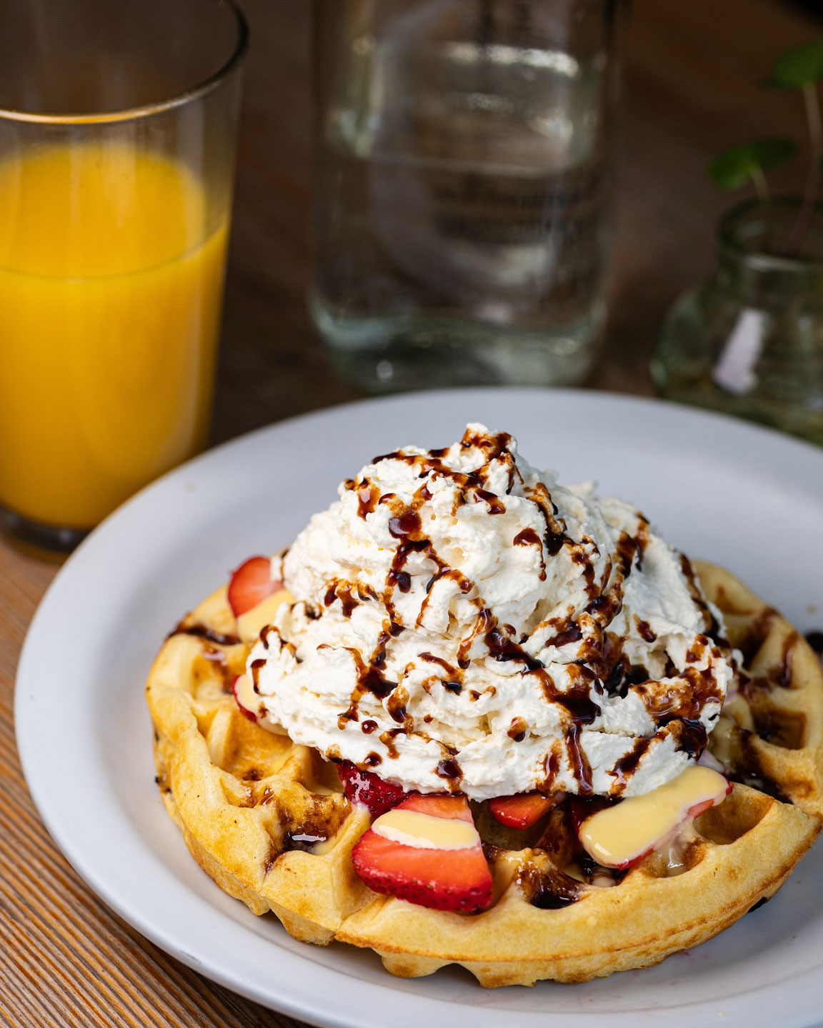 A waffle topped with fresh strawberries, dollops of whipped cream, and drizzled chocolate sauce. Beside it is a glass of orange juice.