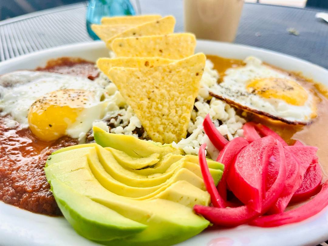 A colorful plate of huevos rancheros features sunny-side-up eggs, sliced avocado, tortilla chips, pickled onions, beans, and melted cheese. A lively, appetizing setting.