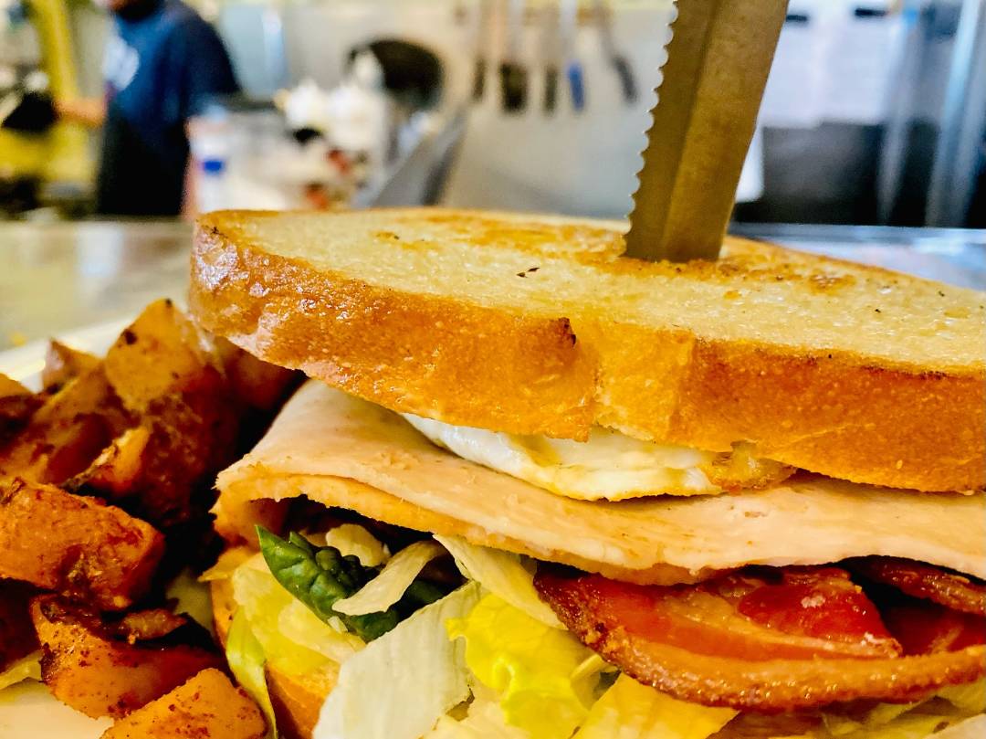 Close-up of a sandwich with turkey, bacon, lettuce, and fried egg between toasted bread, skewered by a knife. Served with crispy potato wedges.
