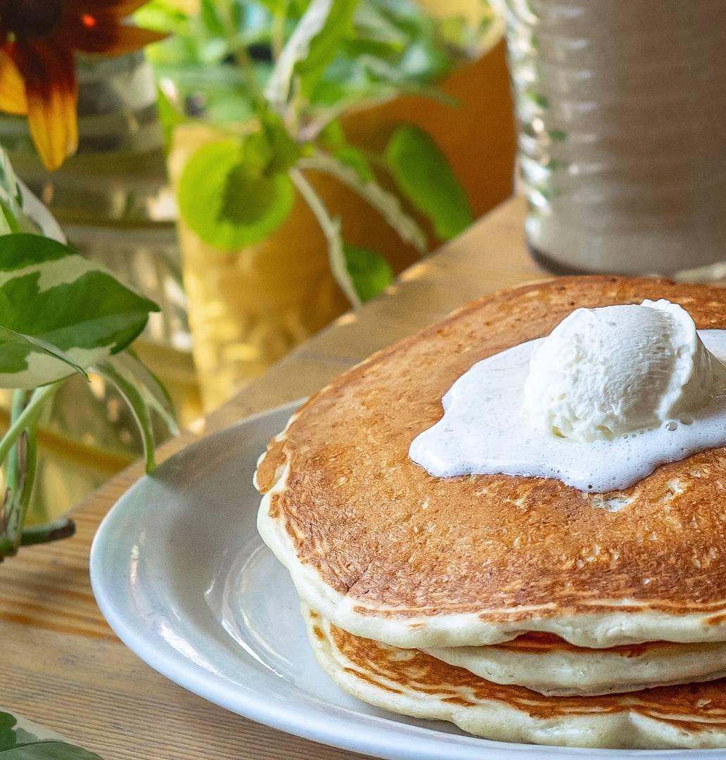 A stack of fluffy pancakes topped with whipped cream on a white plate, set on a wooden table. Lush green plants and soft sunlight create a cozy ambiance.
