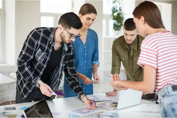 A group of four people are gathered around a table in a bright office, collaborating on documents and using a laptop. Their faces are intentionally blurred for privacy.