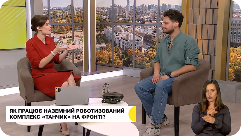 Двоє людей із затуманеними обличчями сидять у студії ток-шоу і розмовляють про роботизовані системи, а третя людина на невеликому вставному екрані перекладає їхню розмову мовою жестів.