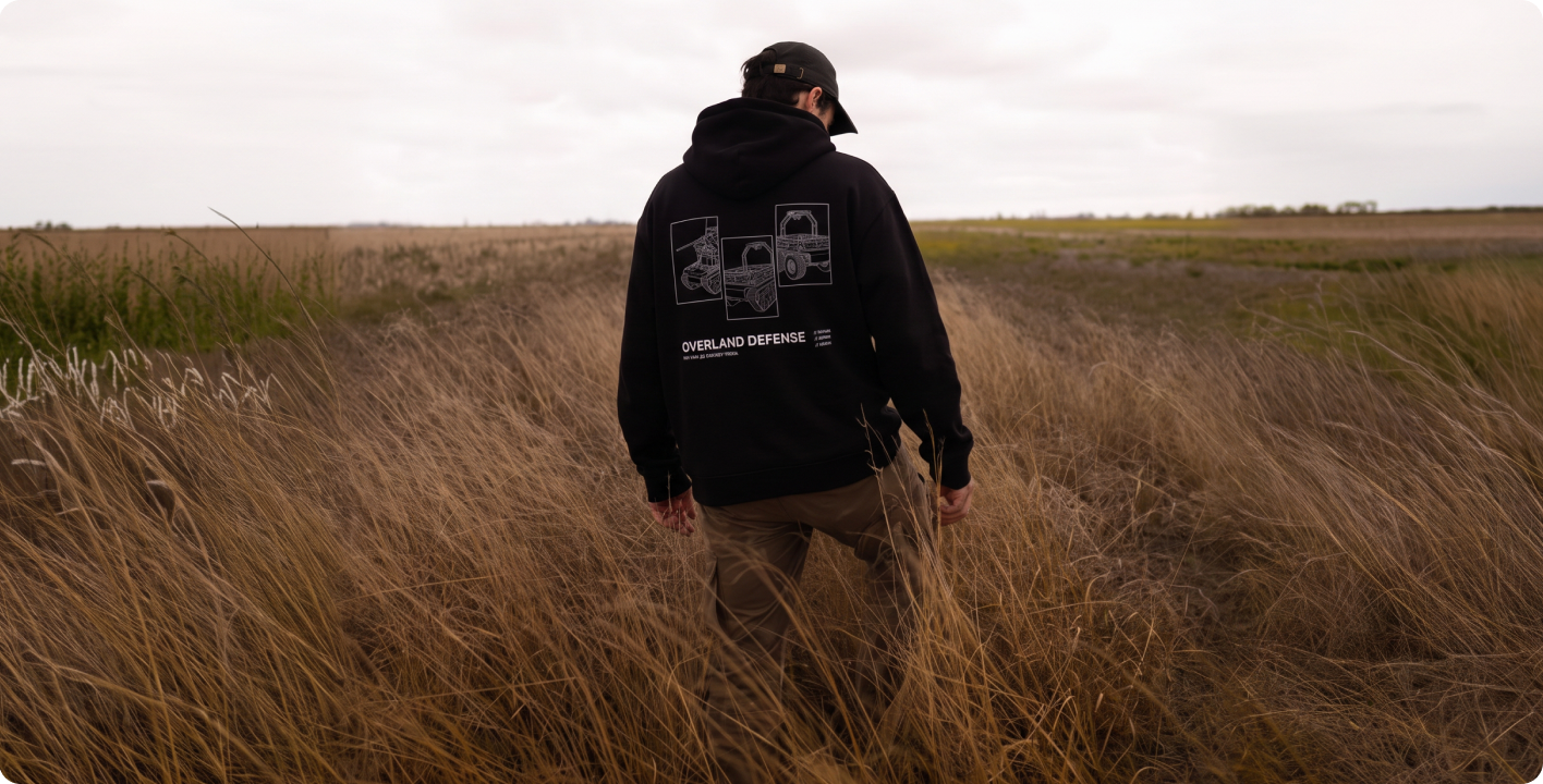 Людина в чорному худі Overland Defense стоїть у полі спиною до камери