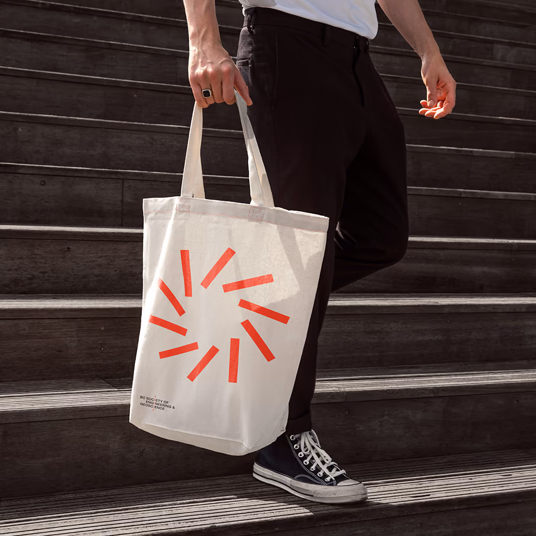 Person wearing black pants and black sneakers with white laces standing on wooden steps, holding a cream-colored tote bag with a red geometric circular design and a small logo near the bottom left corner.