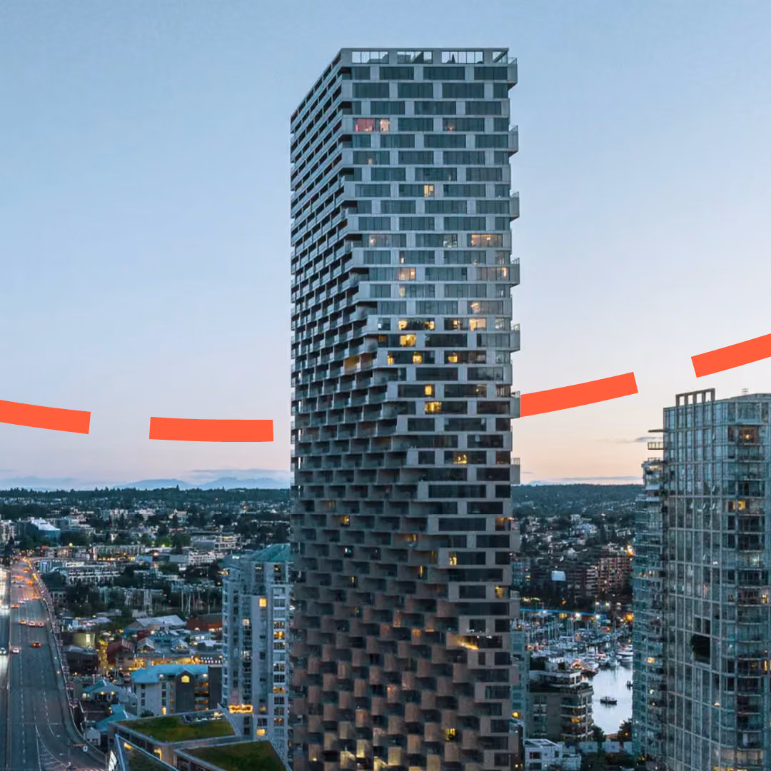 A tall, modern high-rise building with a distinctive blocky, staggered facade illuminated by interior lights, set against a cityscape at dusk with surrounding buildings and distant hills under a clear sky. A dashed red-orange line runs behind it.