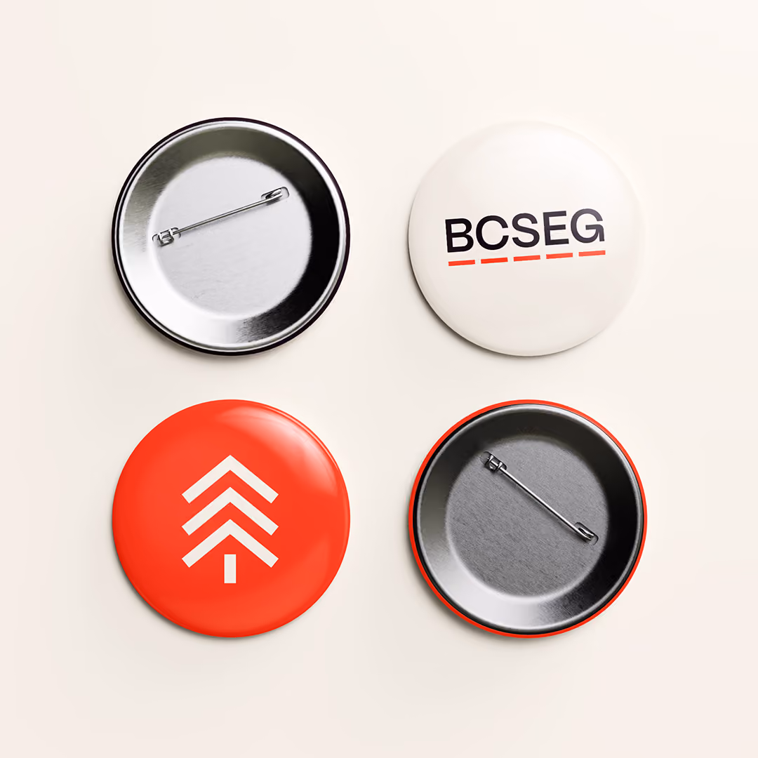 Four round pins arranged in a square on a light background; top left and bottom right pins show metal backs with safety pins, top right pin is white with the dark text 'BCSEG' underlined in red-orange, and bottom left pin is red-orange with a white stylized tree icon on it.