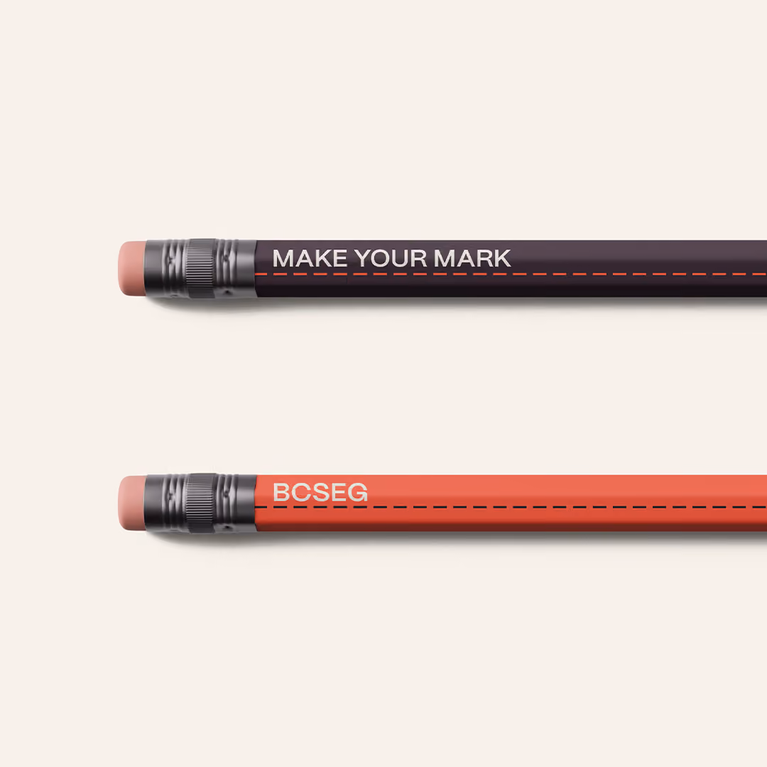 Two pencils laid horizontally on a light surface; the top pencil is black with 'MAKE YOUR MARK' written in white, and the bottom pencil is red with 'BCSEG' written in white, each with an eraser on the left end. Both have a red-orange dashed line running along the pencil.