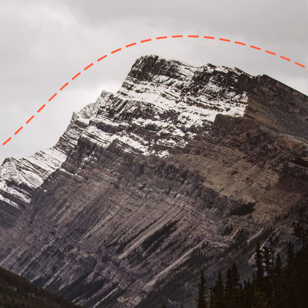 Snow-capped rugged mountain with steep rock layers under a cloudy gray sky, with a dashed red curved line running behing it, with dark pine trees visible at the base.