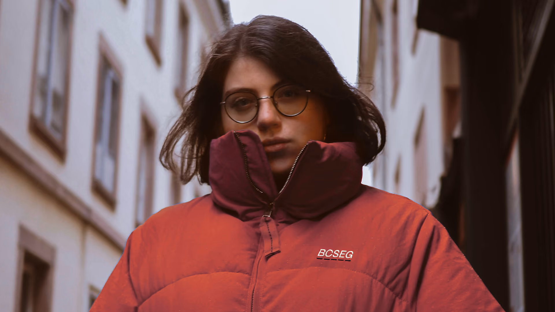 Person with shoulder-length dark hair and round glasses wearing a red puffer jacket with a logo reading BCSEG on it, standing on a city street between buildings.