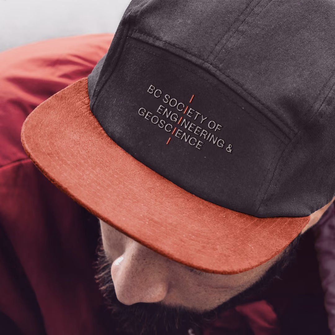 Close-up of a black and rust-colored cap worn by a man with a beard, embroidered with 'BC Society of Engineering & Geoscience' on the front panel.