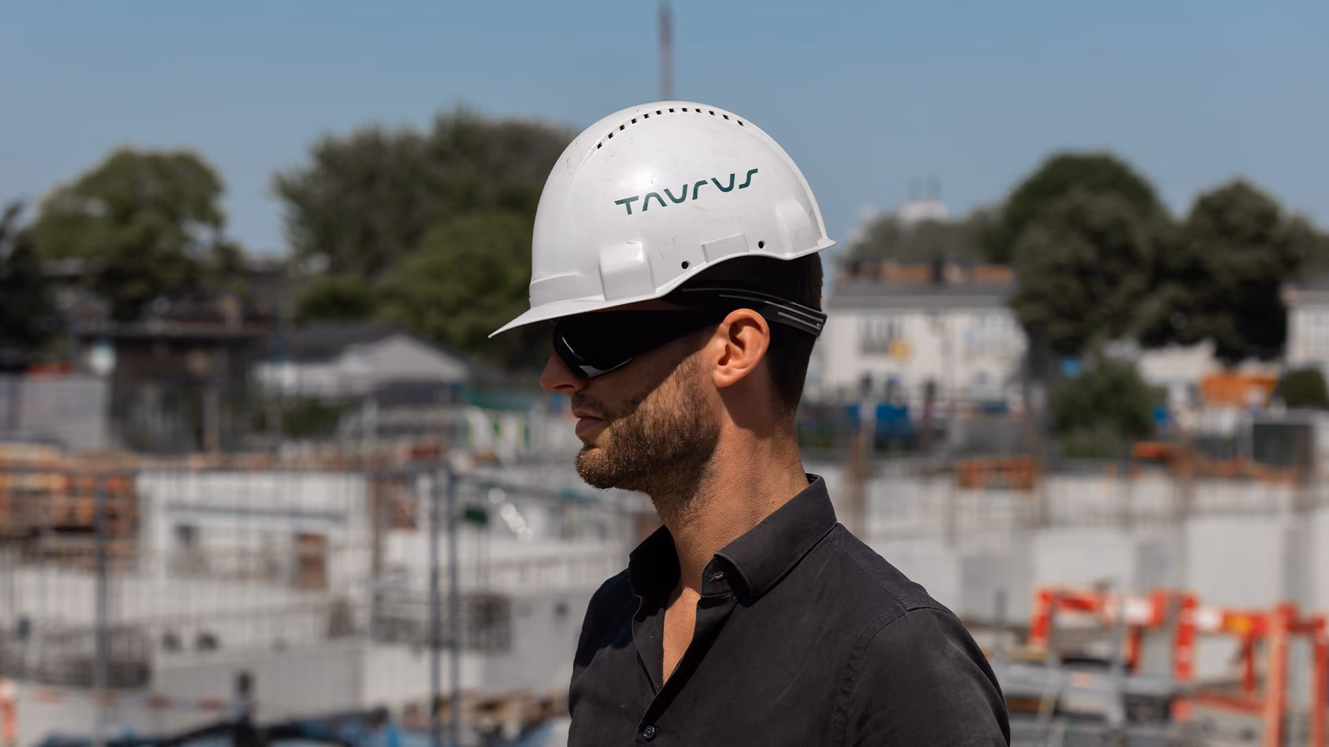 Man in black shirt wearing white hard hat with 'TAURUS' logo and protective sunglasses at a construction site with blurred background.