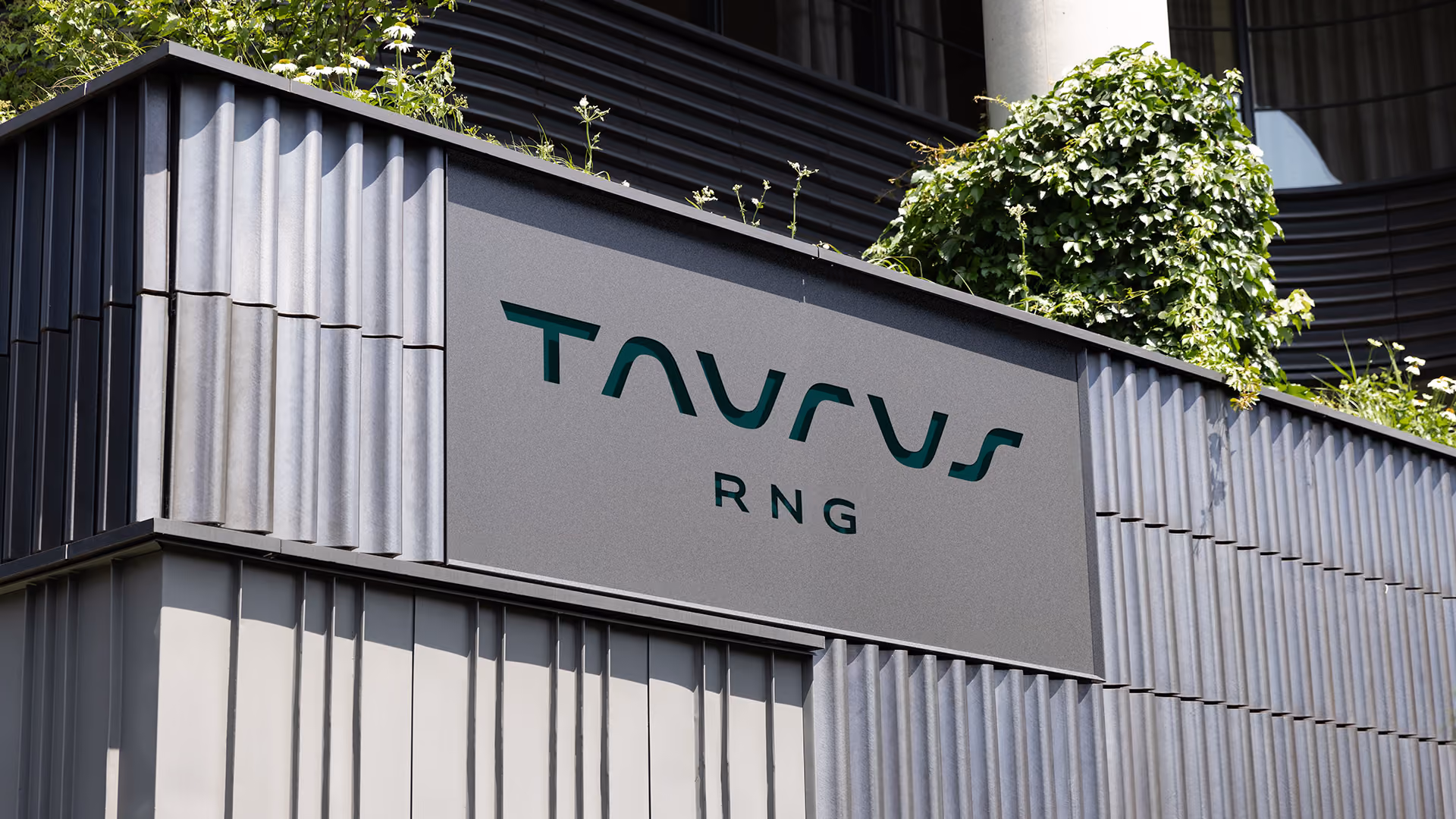 Gray modern building facade with vertical ridges and a sign displaying the word 'TAURUS' in curved stylized letters above the smaller text 'RNG', with green plants growing on top of the structure.