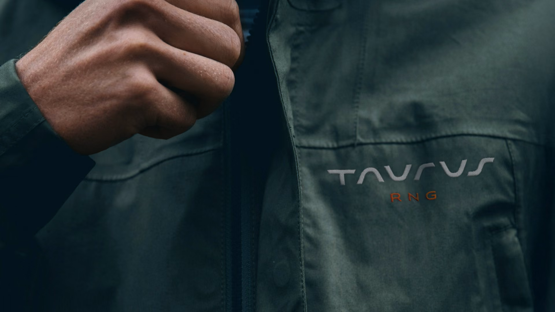 Close-up view of a person's hand adjusting the zipper on a dark green jacket with a modern curved logo reading  'TAURUS RNG' printed on the chest.
