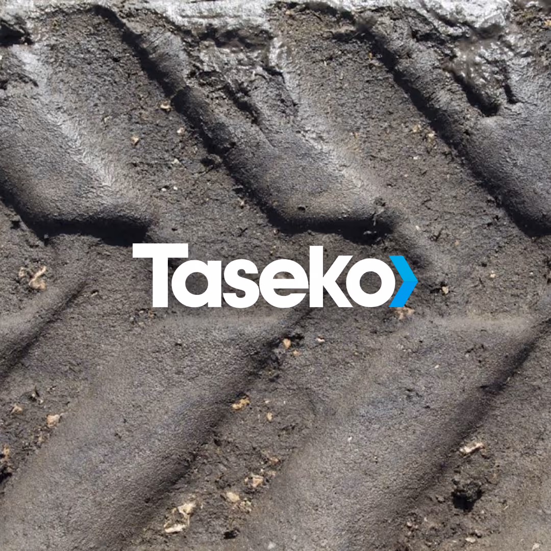 Close-up of a tire tread pattern imprinted in dirt with a logo: the white word 'Taseko' and a blue arrow-like symbol overlaid in the center.