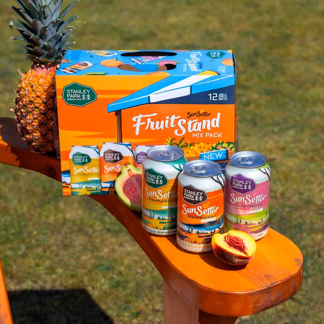 Three Stanley Park Brewing SunSetter wheat ale cans (Pineapple, Peach, Guava flavors) on an orange wooden surface outdoors, with a matching Fruit Stand mix pack box, a whole pineapple, and sliced fruits (guava and peach).