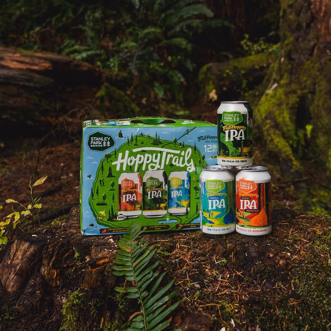 Stanley Park Brewing Hoppy Trails IPA mix pack displayed outdoors on a mossy tree stump, featuring four colorful cans and a green box with a hiking trail illustration.
