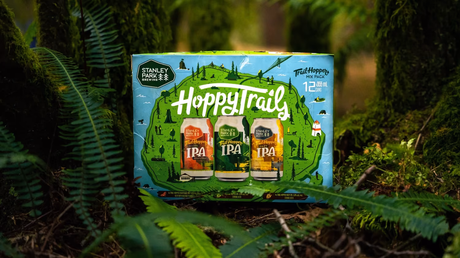 Stanley Park Brewing Hoppy Trails Trail Hoppy Mix Pack 12 cans of IPA displayed outdoors among green ferns and moss-covered trees.