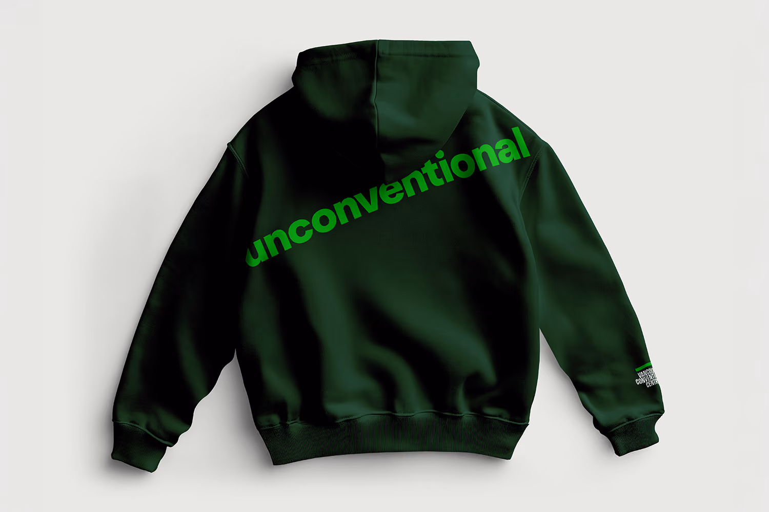 Dark green hoodie laid flat showing the back with the word 'unconventional' printed diagonally in bright green letters, and a small logo on the right sleeve cuff.