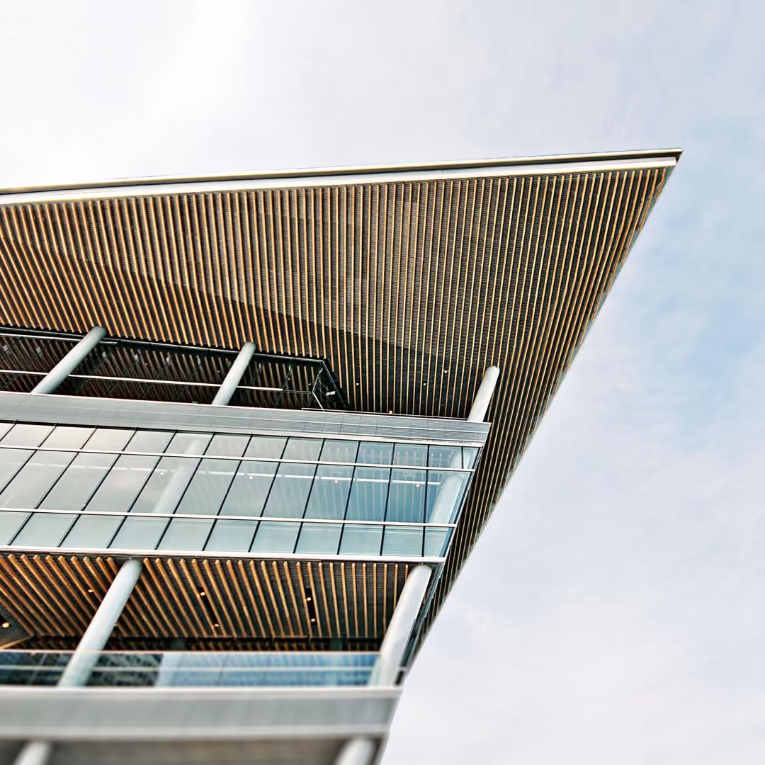 Upward view of a corner of the Vancouver Convention Centre, a modern building with a sharp overhanging roof, vertical pillars, and large glass windows under a light blue sky.