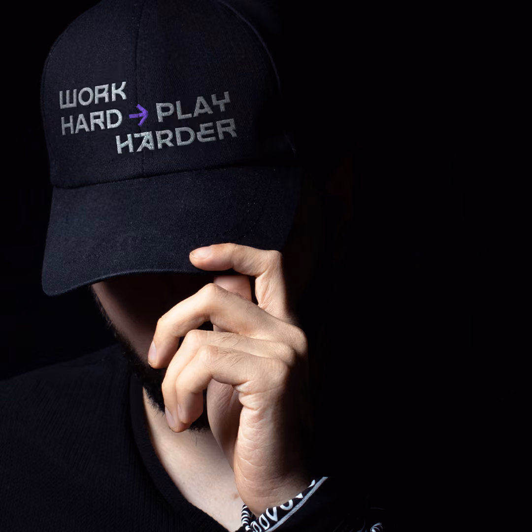 Close-up of a person wearing a black cap with the embroidered text 'WORK HARD → PLAY HARDER' in custom type in gray and purple on the front, with the person's hand adjusting the cap brim against a black background.