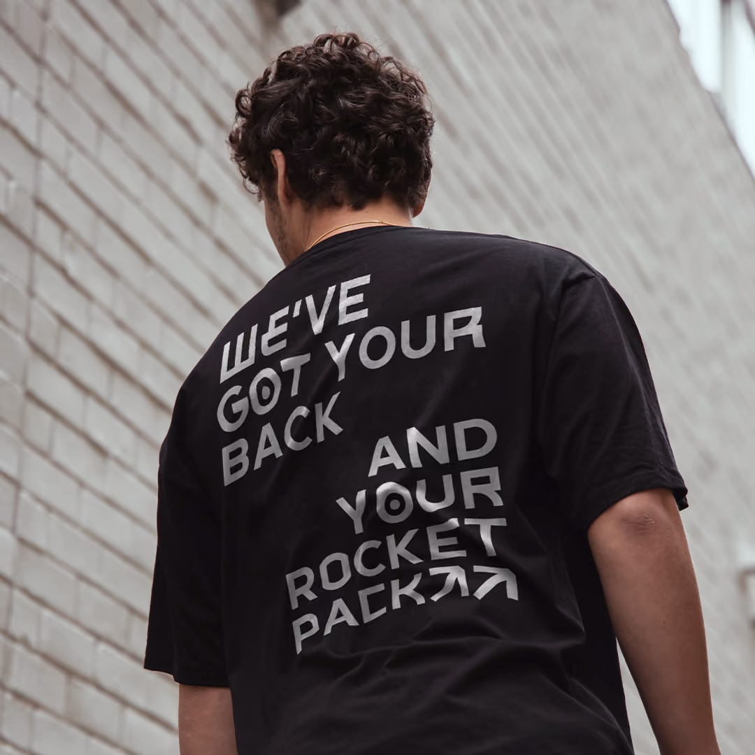 Person with curly hair wearing a black t-shirt with futuristic text on the back that reads, 'WE'VE GOT YOUR BACK AND YOUR ROCKET PACK' in stylized white letters, standing against a light gray brick wall.
