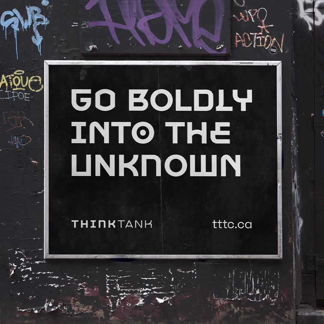 A black sign with bold white text reading 'GO BOLDLY INTO THE UNKNOWN' in a stylized font, with 'THINK TANK' and 'tttc.ca' below it, mounted on a black wall covered in colourful graffiti.
