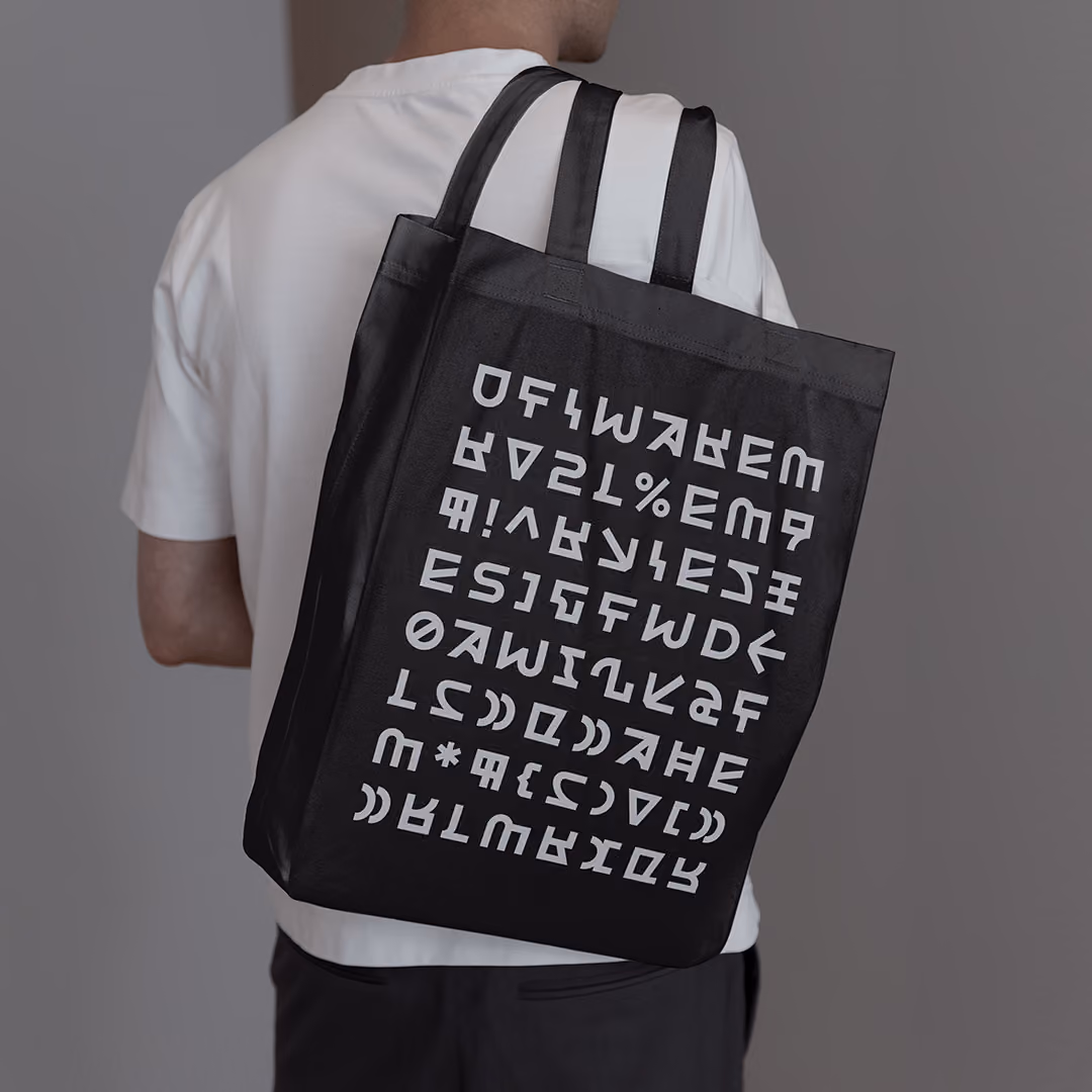 Person wearing a plain white t-shirt carrying a black tote bag with two handles, featuring a grid of graphic white stylized geometric letters and symbols on the front.