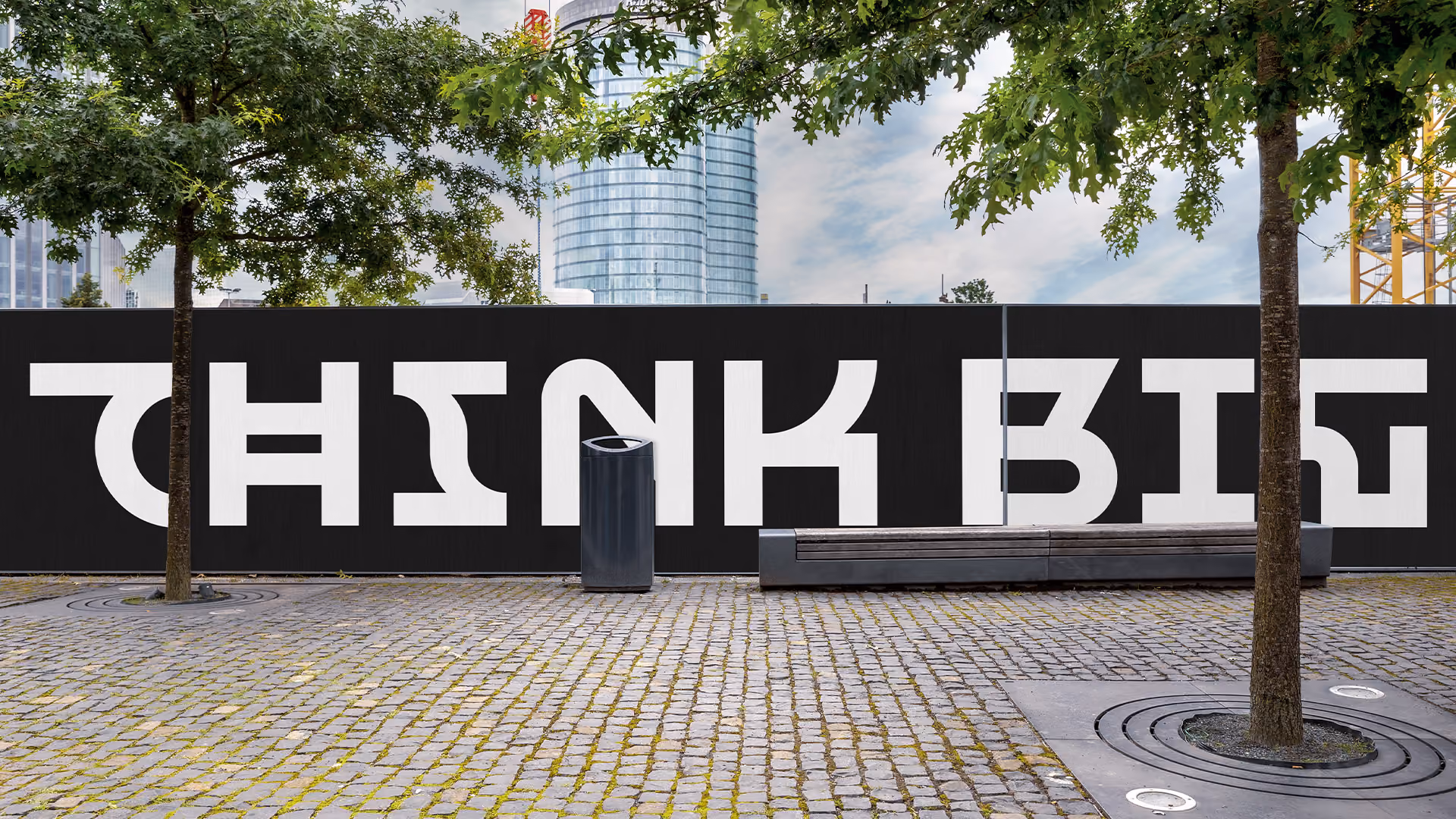 Outdoor urban scene with cobblestone pavement, trees, a trash bin, and a long bench in front of a black wall featuring large white stylized text that reads 'THINK BIG' in the shifting reality typeface.