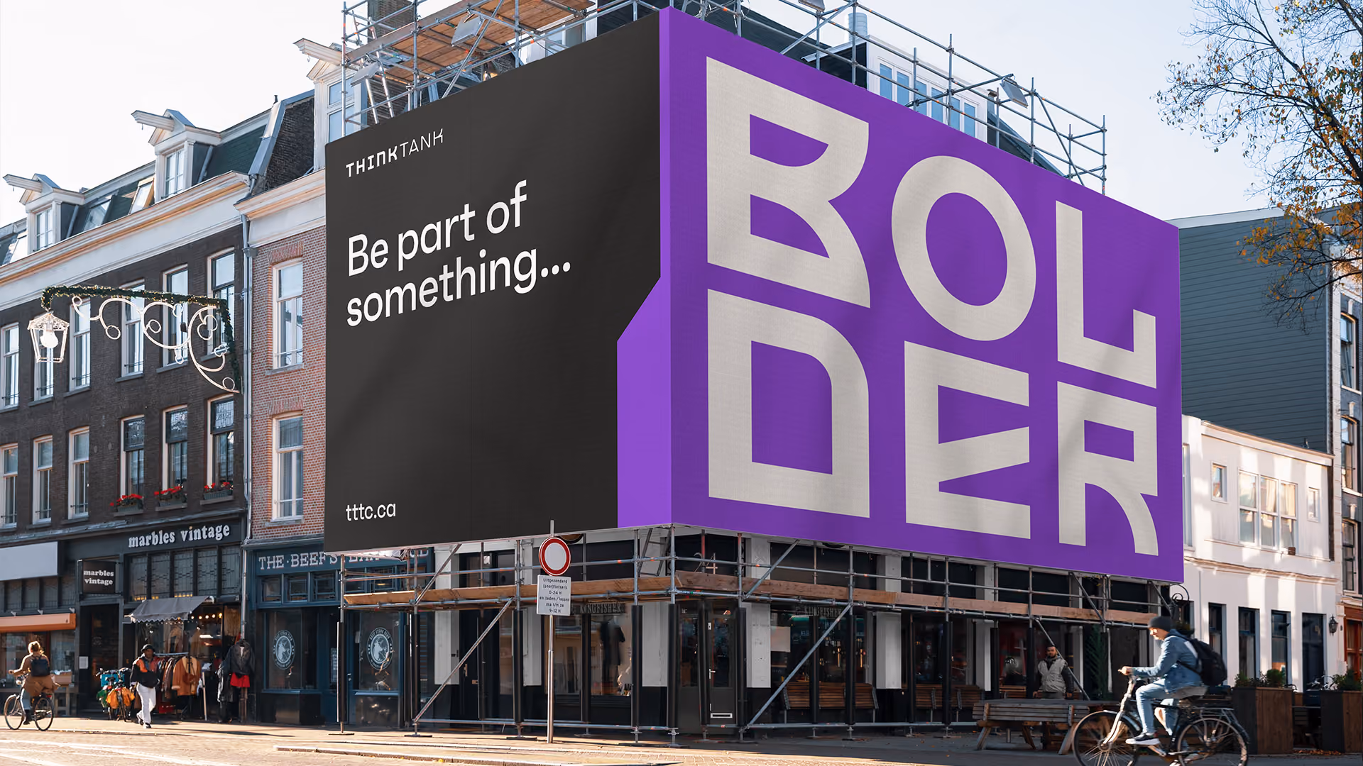 Large corner billboard on building scaffolding in a city street displays text 'Be part of something...' and 'BOLDER' in bold white letters on black and purple backgrounds.