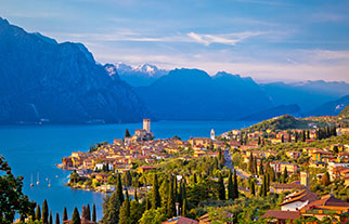 Scenic lake town with historic buildings and a castle, surrounded by green trees and mountains under a blue sky.