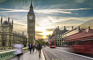 Blurred red double-decker buses and pedestrians on Westminster Bridge with Big Ben and the Houses of Parliament at sunset in London.