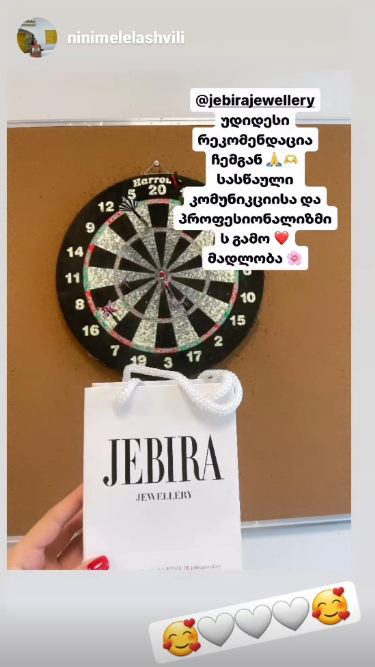 Hand holding a white JEBIRA Jewellery shopping bag in front of a dartboard mounted on a corkboard.